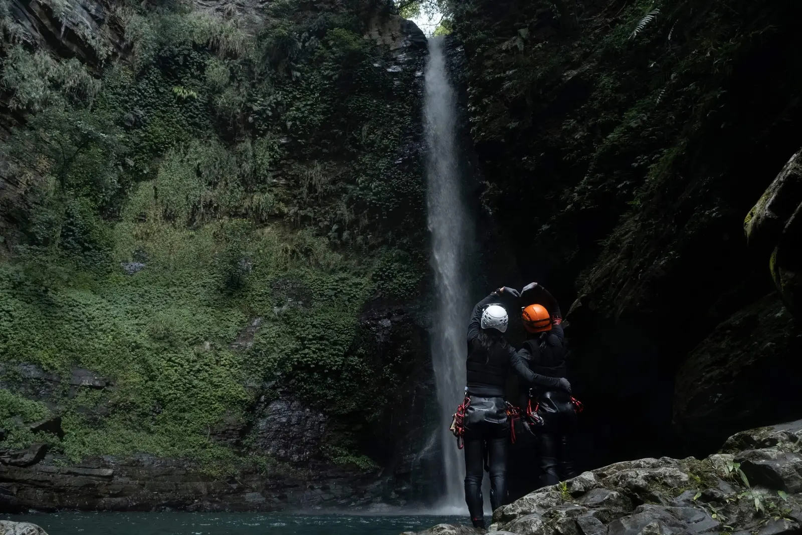 Two people making a heart shape in front of a tall waterfall in a jungle.